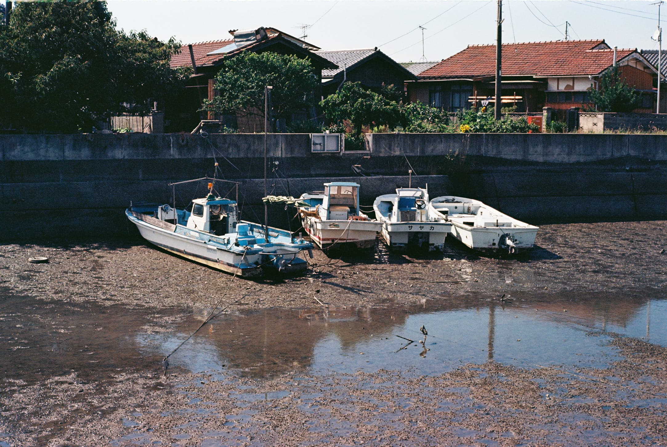 three boats on the mud beside a pool reflecting the sky, in front of tile-roofed houses